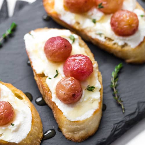 Image of roasted grape crostini on a black slate board, topped with ricotta, roasted grapes, and a drizzle of honey.