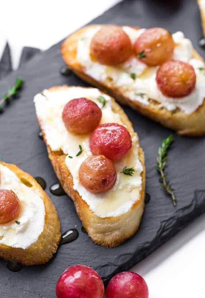 Image of crostini on a black slate background. They are topped with ricotta cheese, honey, and roasted grapes. 