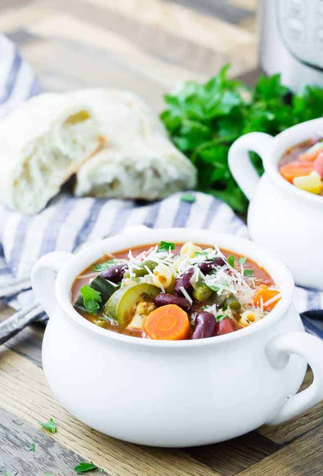 Image of pressure cooker minestrone in a bowl. Another bowl, parsley, bread, and an Instant Pot are in the background.