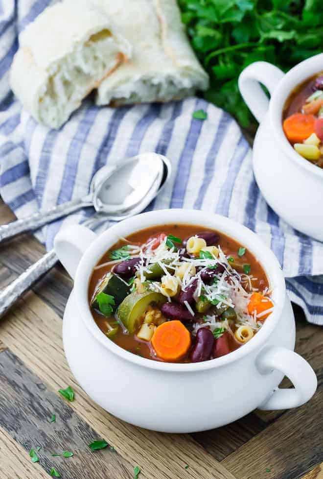 Image of healthy minestrone soup in a white bowl with another bowl in the background as well as bread, parsley, and two spoons.