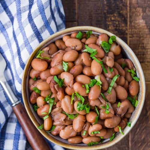 Image of pressure cooked pinto beans in a small bowl on a wooden background. A fork and a blue and white checkered cloth are also pictured.