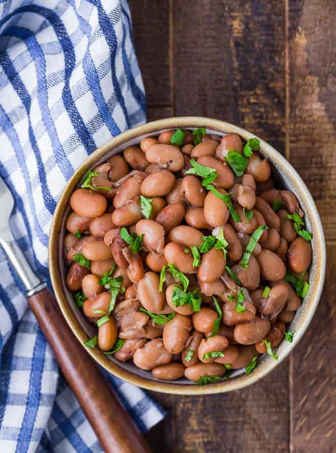 Image of pressure cooked pinto beans in a small bowl on a wooden background. A fork and a blue and white checkered cloth are also pictured.