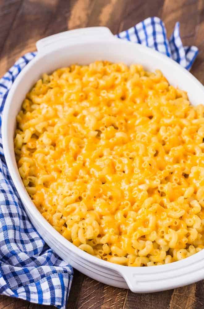 Baked mac and cheese in a white oval casserole dish next to a blue and white checkered linen on a wooden background.