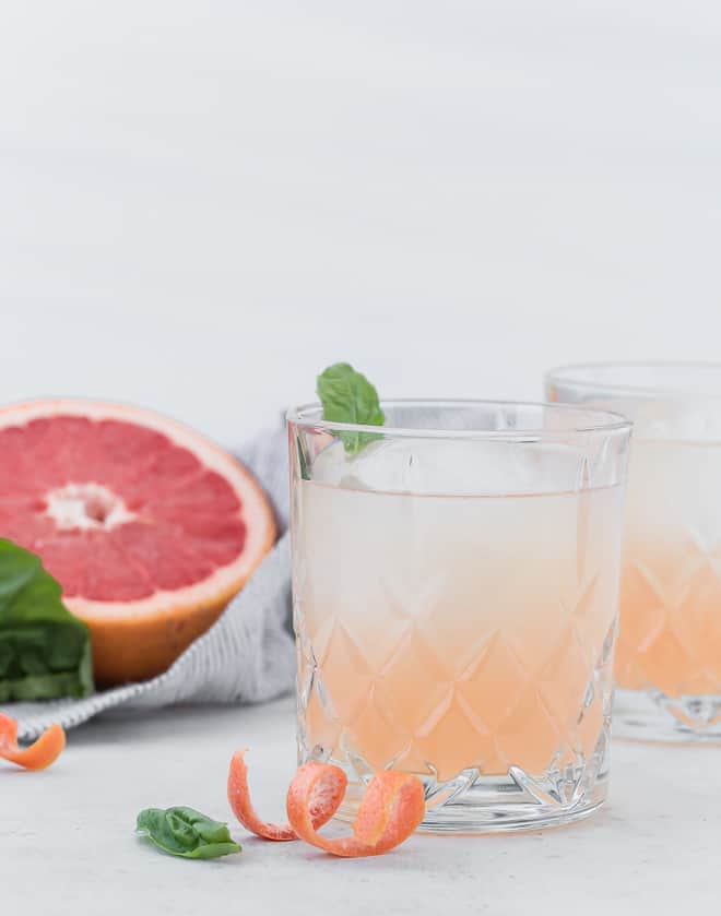 Image of a pale pink grapefruit cocktail with a half of a grapefruit in the background, also basil leaves are pictured.