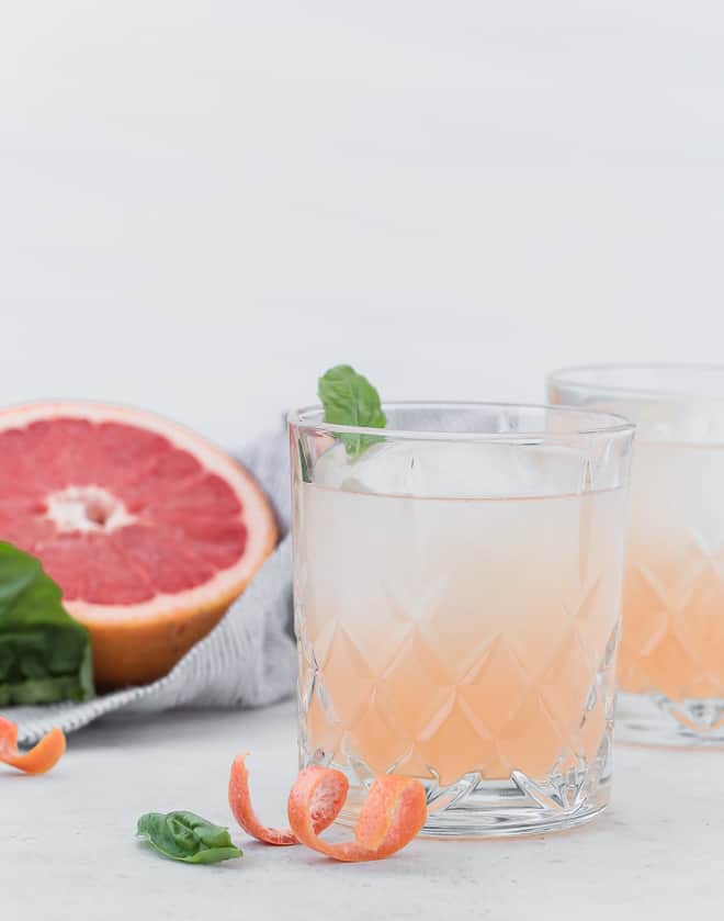 Image of a pale pink grapefruit cocktail with a half of a grapefruit in the background, also basil leaves are pictured. 