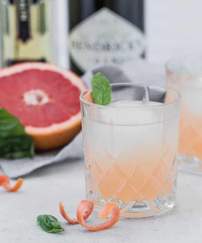 Image of a pale pink cocktail, garnished with a fresh basil leaf. Hendricks Gin and elderflower liqueur are pictured in the background. A curl of grapefruit rind is in the foreground of the photograph.