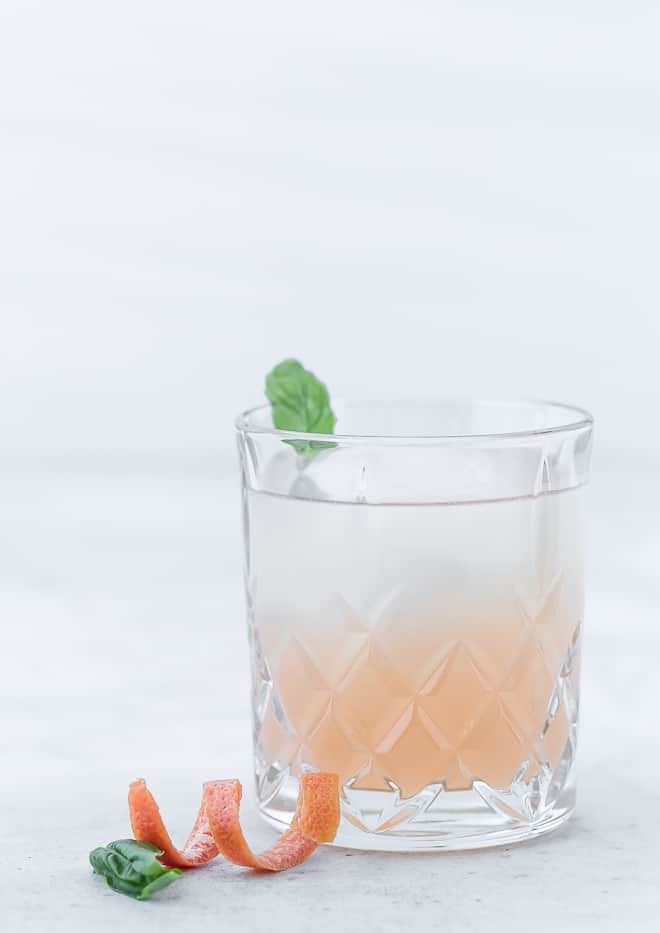 Image of a pink grapefruit cocktail made with gin and elderflower liqueur. The drink is garnished with a fresh basil leaf.