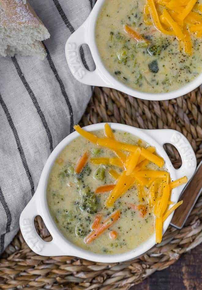 Image of two bowls of broccoli cheddar soup in white crocks with handles. Soup is garnished with additional cheddar cheese.