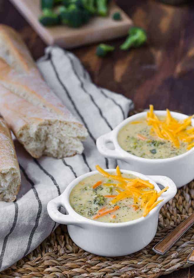 Image of two bowls of instant pot broccoli cheese soup, with fresh broccoli and bread in the background of the image.