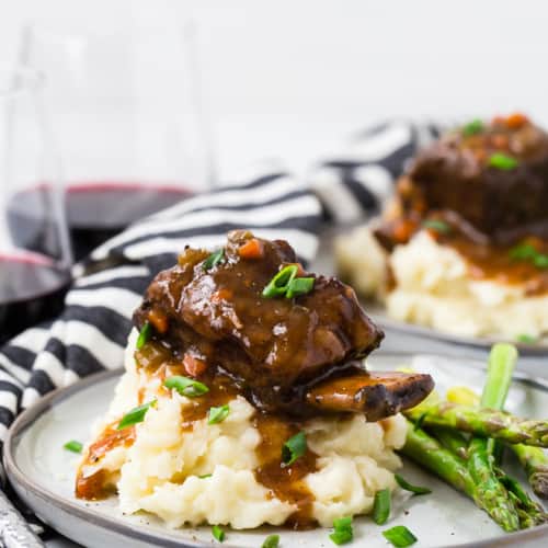 Image of a short rib on top of a pile of creamy mashed potatoes with brown gravy dripping down them. Everything is garnished with fresh chives.