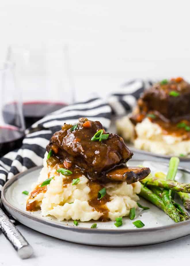 Image of a short rib on top of a pile of creamy mashed potatoes with brown gravy dripping down them. Everything is garnished with fresh chives.