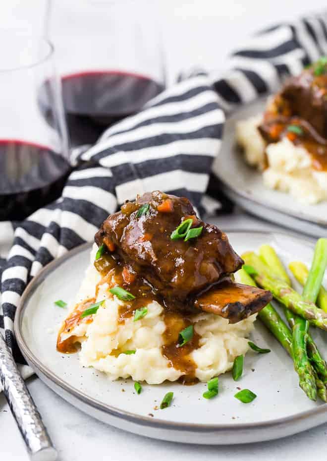 Image of Instant Pot short ribs on a pile of mashed potatoes, garnished with fresh chives. Short ribs are served with sauteed asparagus and a glass of red wine.