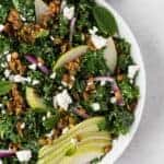 Image of a kale salad in a white bowl topped with pears, feta cheese, red onions, and savory granola.