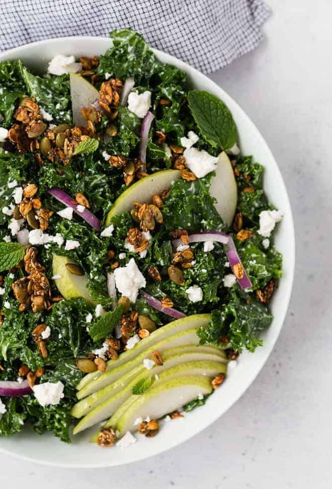 Image of a kale salad in a white bowl topped with pears, feta cheese, red onions, and savory granola. 