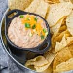 Image of mexicali dip in a black bowl with two handles. Dip is surrounded by chips on a tray.