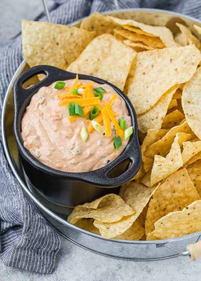 Image of mexicali dip in a black bowl with two handles. Dip is surrounded by chips on a tray.