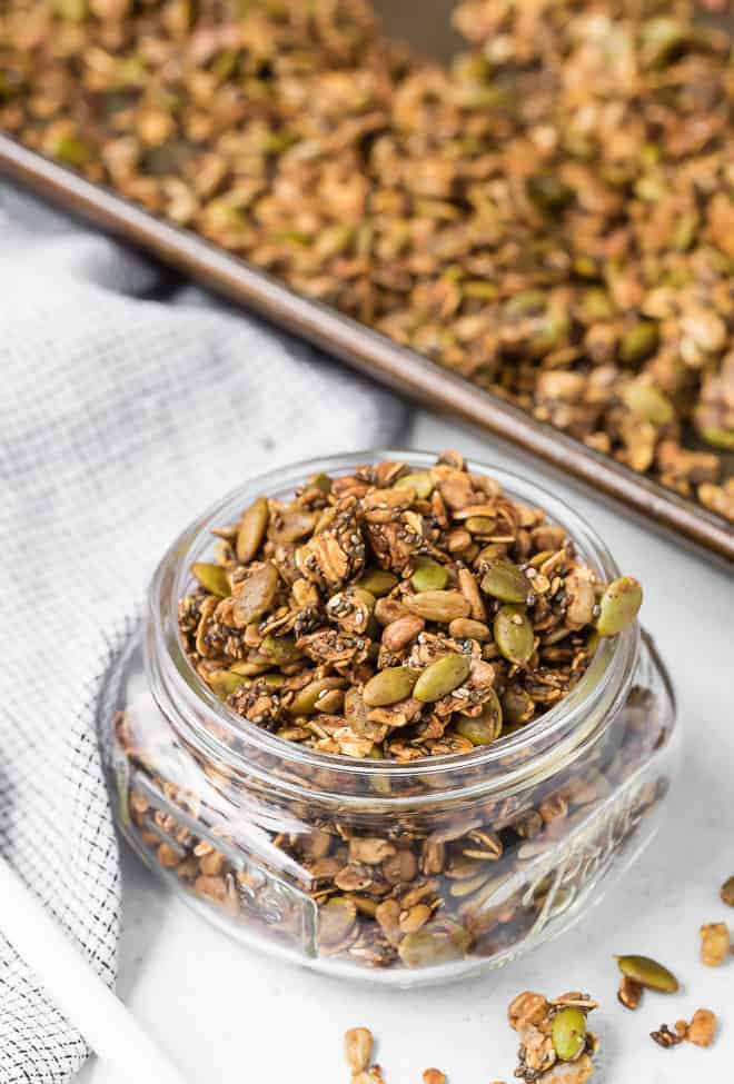 Image of savory granola in a jar, spilling out. A pan of granola is also pictured in the background.