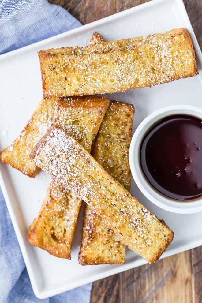 Image of a square white plate with 5 french toast sticks on it, sprinkled with powdered sugar. Also pictured is a small glass bowl of maple syrup.