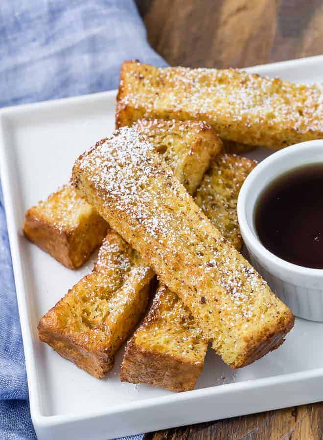 Air fryer french toast sticks on a white plate with a bowl of maple syrup for dipping.