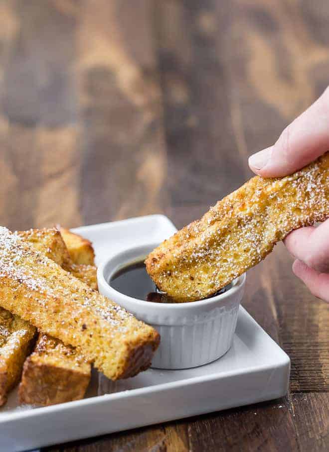 A hand dipping a French toast stick into a bowl of maple syrup on a plate, next to more French toast sticks.