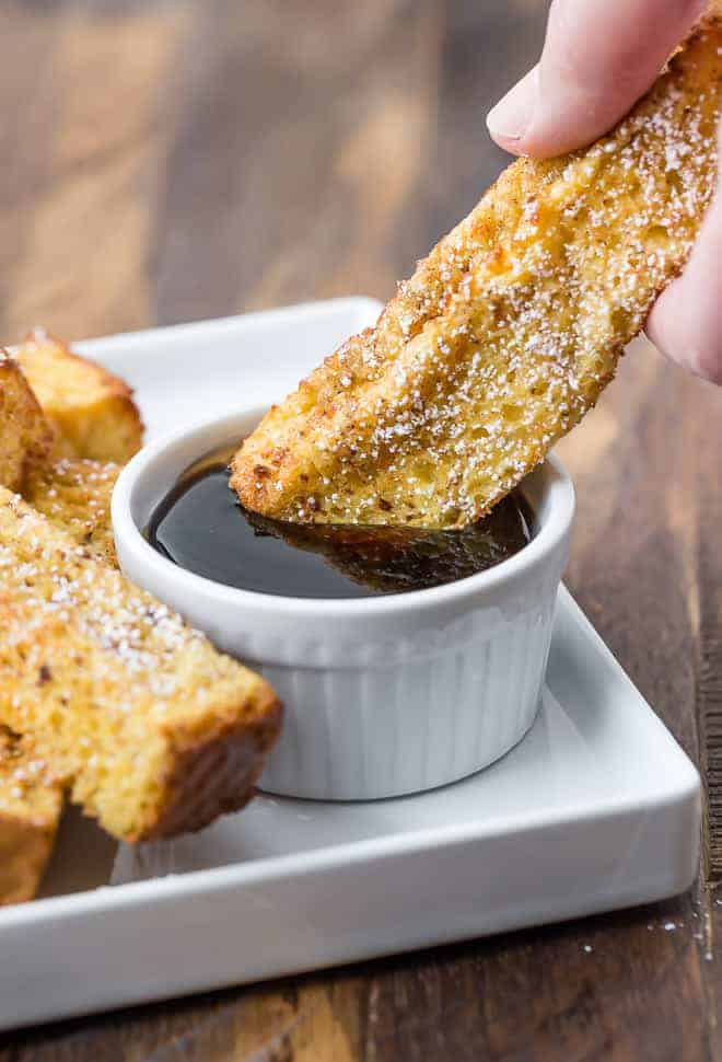 A hand dipping a French toast stick into a bowl of maple syrup on a plate, next to more French toast sticks.