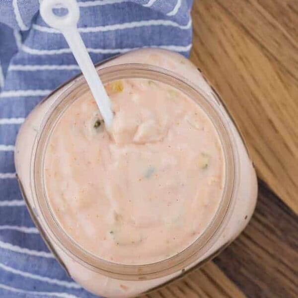 Overhead image of homemade thousand island dressing in a small jar with a little white spoon in it. Jar is placed on a wooden surface with a blue and white towel.