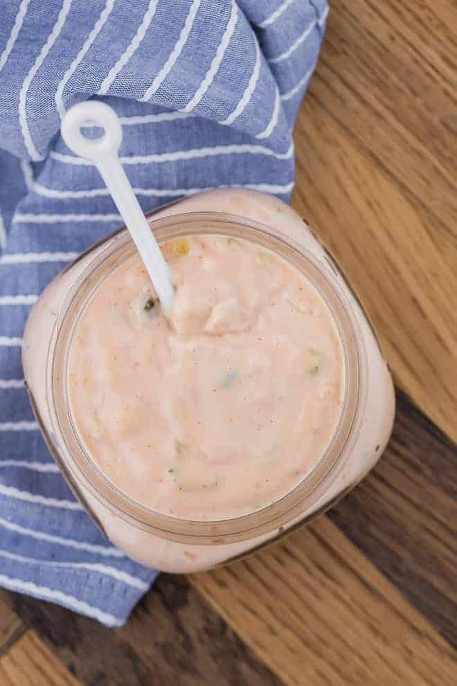 Overhead image of homemade thousand island dressing in a small jar with a little white spoon in it. Jar is placed on a wooden surface with a blue and white towel.