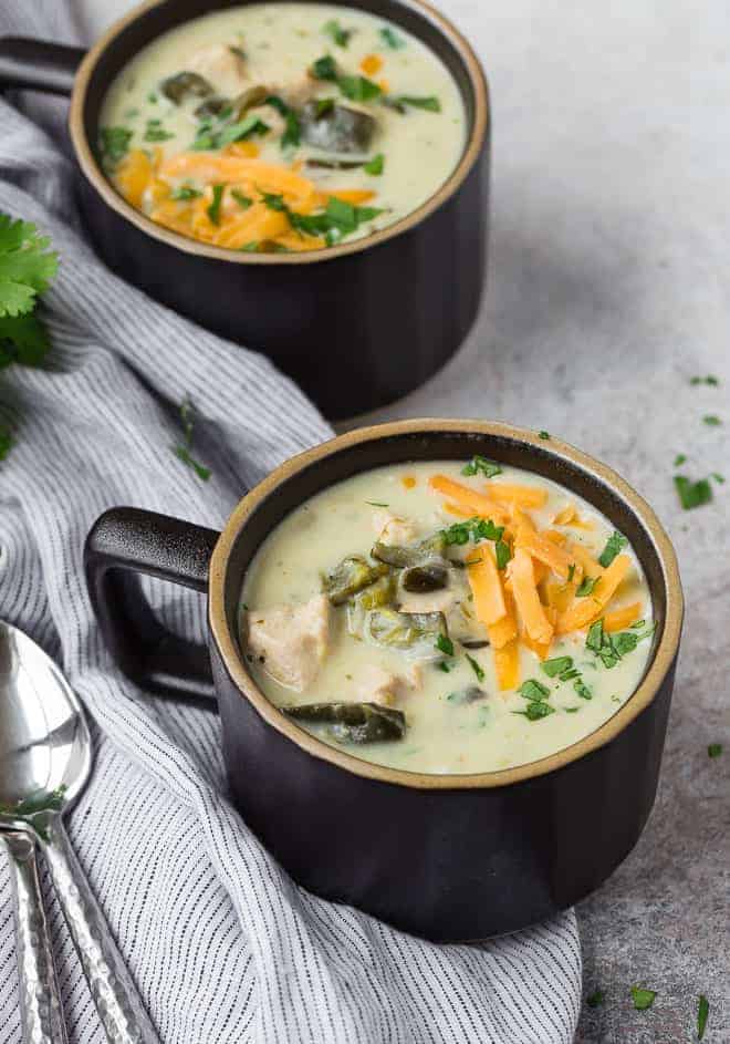 Creamy soup in two black mugs, topped with cheddar cheese and cilantro.