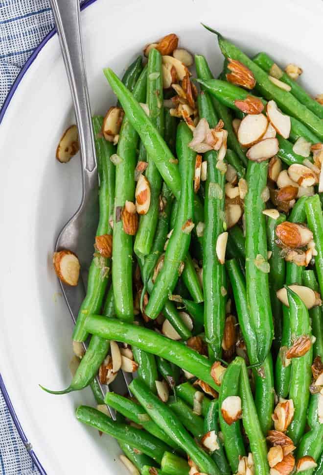 Image of green beans with lemon zest and almonds on a white and blue plate, with a serving utensil also pictured.
