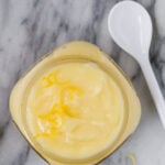 Image of pale yellow lemon cake filling in a small jar with a white spoon next to it.