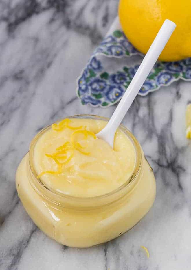 Image of lemon cake filling in a small jar with a white spoon. A lemon is pictured in the background.