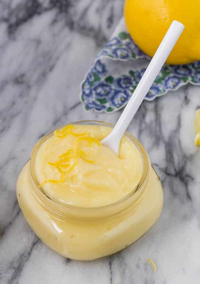 Image of lemon cake filling in a small jar with a white spoon. A lemon is pictured in the background. 