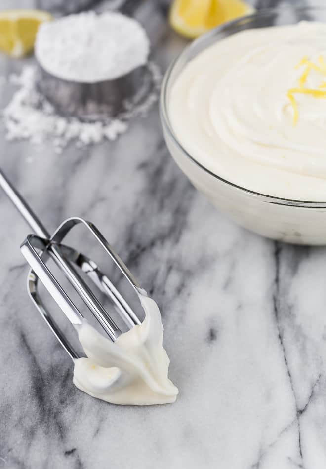 Image of lemon cream cheese frosting in a glass bowl. A beater is next to it, with frosting on it. In the background are powdered sugar and lemons.