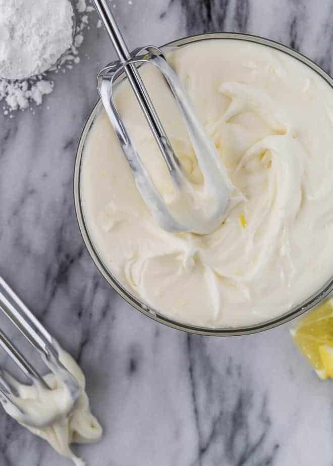 Image of cream cheese frosting with two beaters pictured. Lemons and powdered sugar are also pictured. 