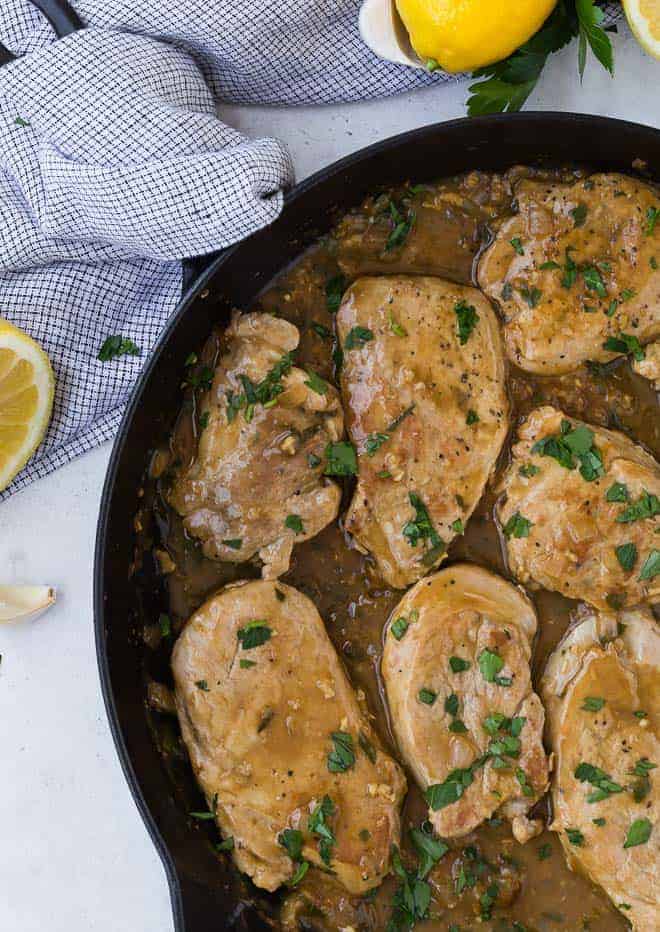 Image of pork tenderloin slices cooked in a lemon garlic sauce, in a black frying pan. Lemons, garlic and fresh parsley are sprinkled throughout the photo.,