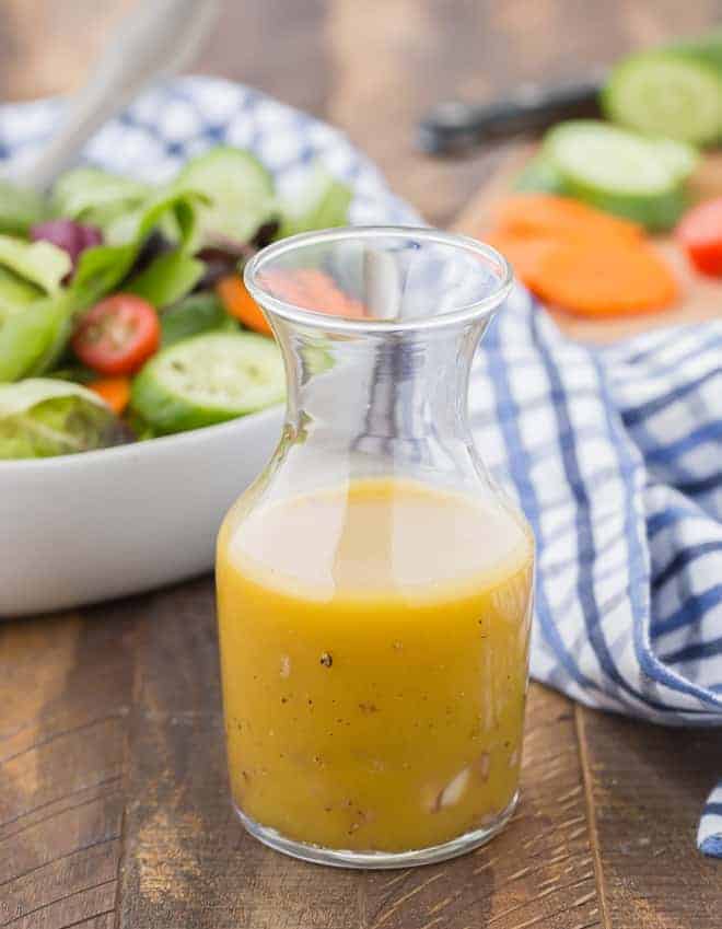 Image of red wine vinaigrette in a small jar. A tossed salad and chopped vegetables are pictured in the background.