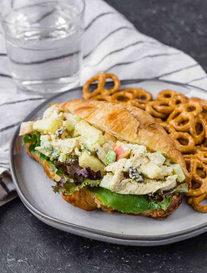 Image of chicken salad made with curry on a croissant with greens. Mini pretzels are also on the plate.