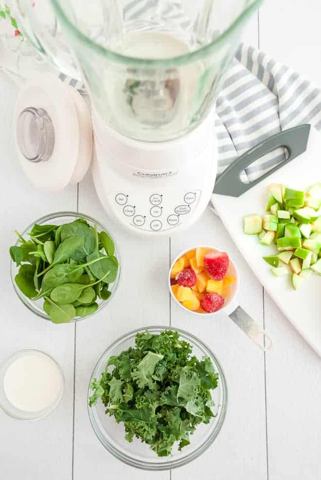 Overhead view of an empty blender, surrounded by bowls of spinach, kale, tropical fruit, and green apples.
