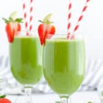 Two green drinks in stemmed glasses with red and white paper straws. Strawberries garnish the edge of the glasses.