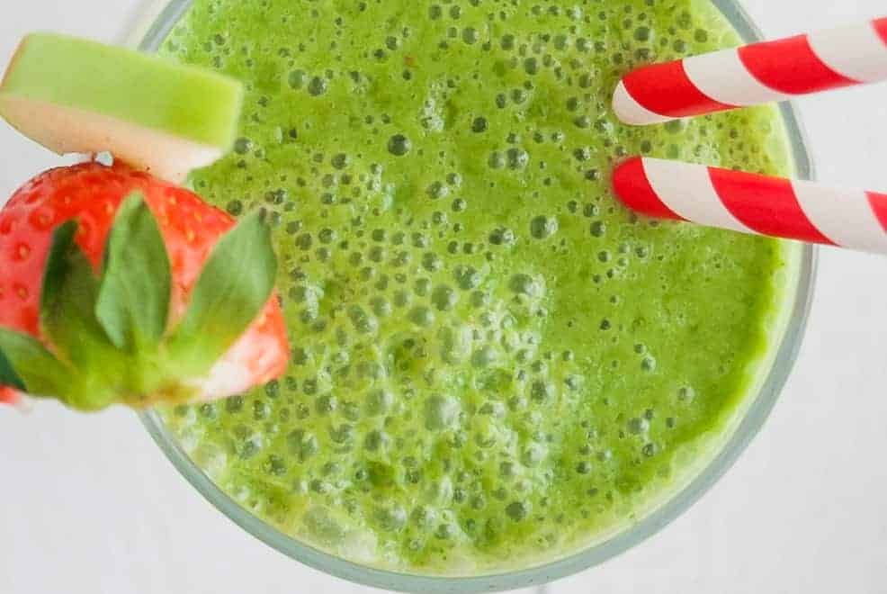 Overhead view of a green smoothie in a glass, garnished with a red strawberry and a slice of green apple.