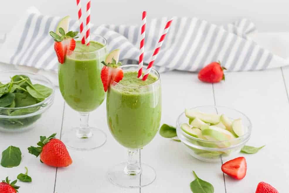 Two green smoothies in stemmed glasses with red and white straws. Also pictured are strawberries, green apples, and a striped linen. 