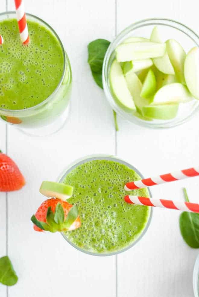 Overhead view of two green drinks with red and white straws. A bowl of cut up green apples are also pictured.