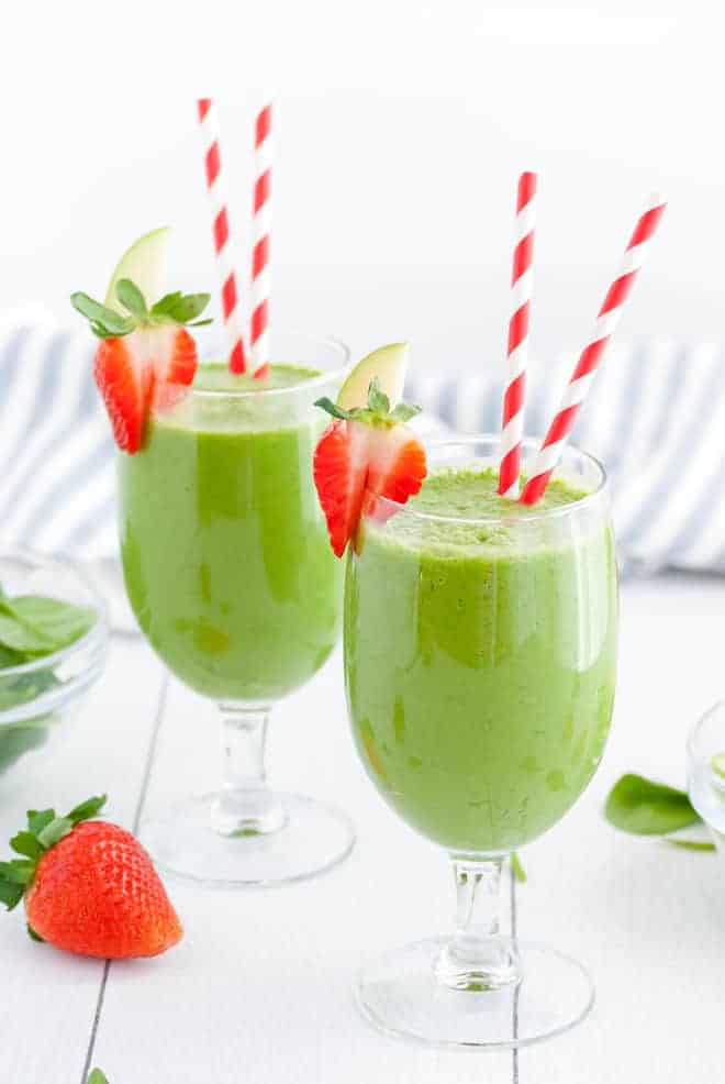 Two stemmed glasses, each with two red and white striped straws. Each glass is filled with green liquid and topped with fresh strawberries.