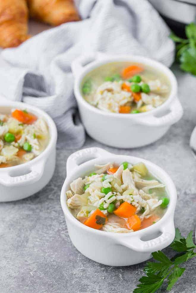 Three small white bowls of soup containing chicken, rice, and vegetables. Croissants are pictured in the background.