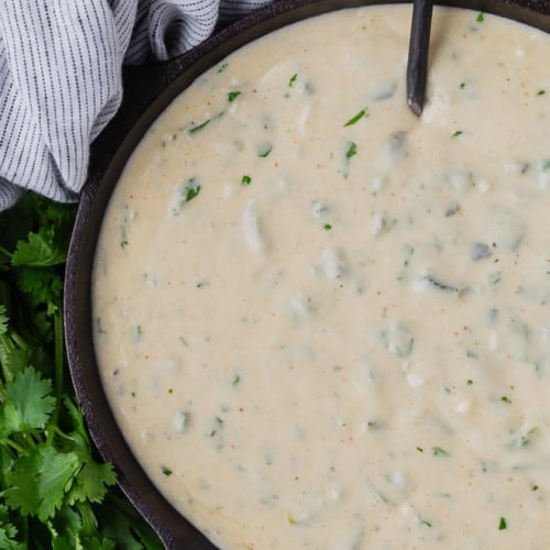 Queso dip in a black pan with a black spoon, next to a black and white linen, and a bunch of fresh cilantro.
