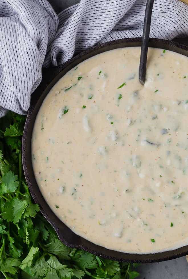 Queso dip in a black pan with a black spoon, next to a black and white linen, and a bunch of fresh cilantro. 