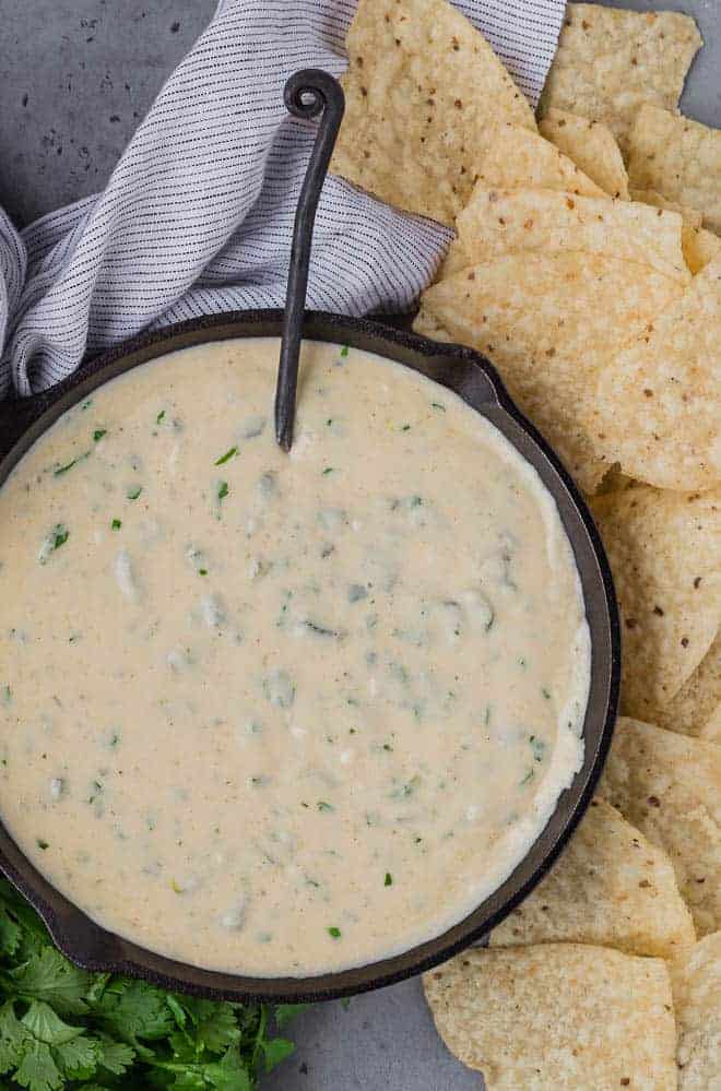 Overhead view of poblano queso in a black skillet, with a black spoon. Also pictured are chips, a linen, and fresh cilantro. 