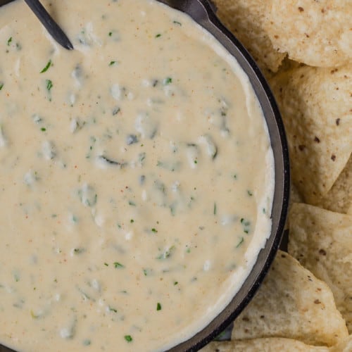 White queso dip in a black skillet, with a black spoon. Queso is studded with green poblano peppers and fresh cilantro, and placed next to tortilla chips.
