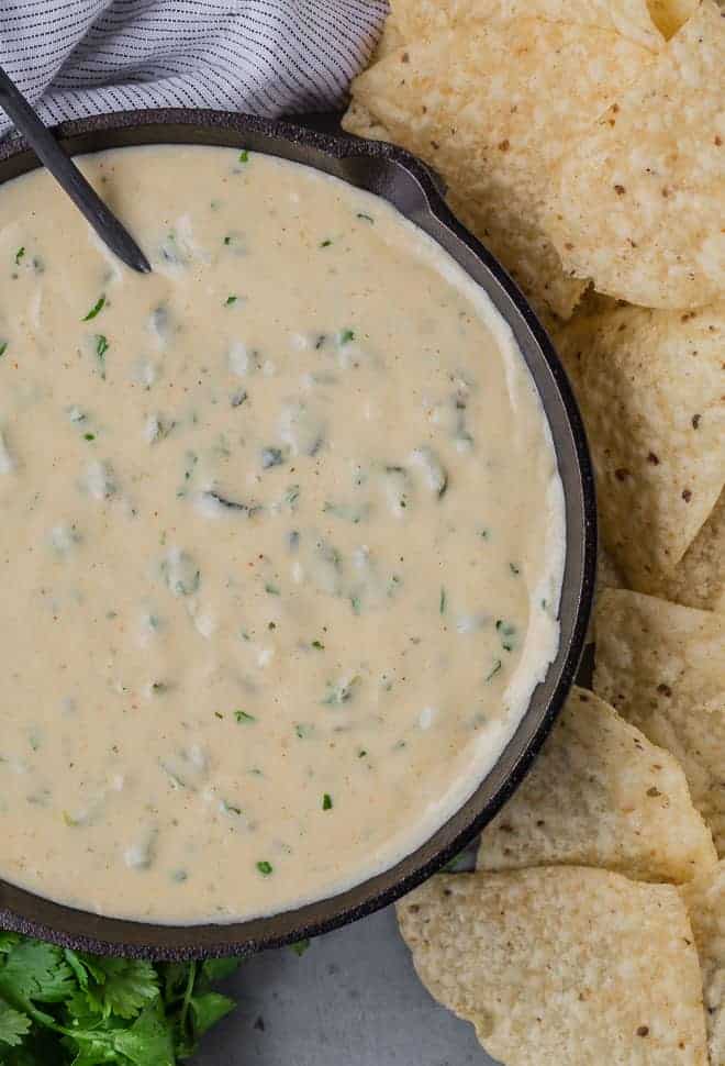 White queso dip in a black skillet, with a black spoon. Queso is studded with green poblano peppers and fresh cilantro, and placed next to tortilla chips.