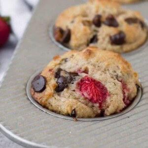 Close-up image of a muffin flecked with fresh strawberries and gooey dark chocolate chips. More strawberries and chocolate chips are pictured in the background.